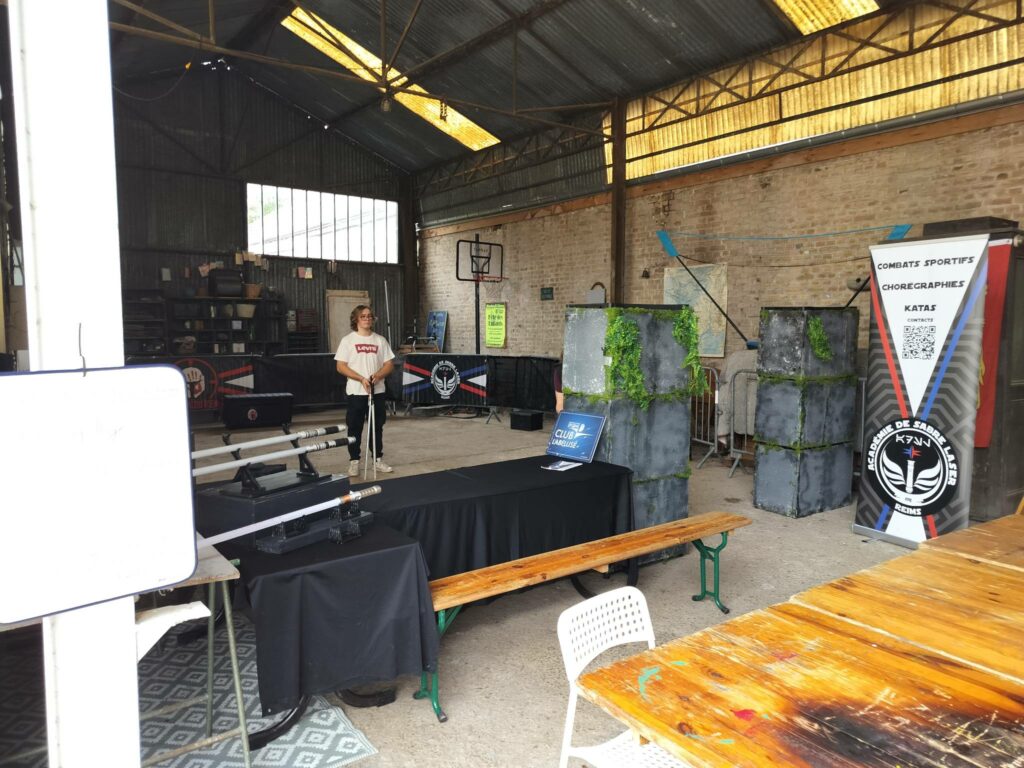 Jeune membre de l'ASL Reims présentant des sabres laser sur le stand de l'Académie de Sabre Laser de Reims à la 25e Fête des Enfants de Nanteuil-sur-Aisne. Le stand expose plusieurs répliques de sabres laser sur une table noire, avec des bannières du club et des décors de combat en arrière-plan dans un grand hall.